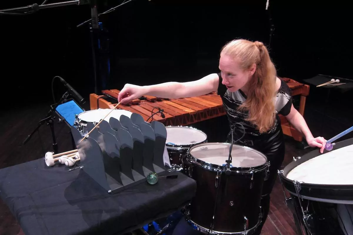 a photo of Bonnie Whiting in performance, scraping a metal mail organizer with a wooden stick, in the middle of a large percussion setup