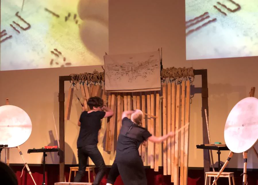 Bonnie Whiting and Jennifer Torrence in performance, reading a graphic score, playing a tall instrument built of large wooden sticks
